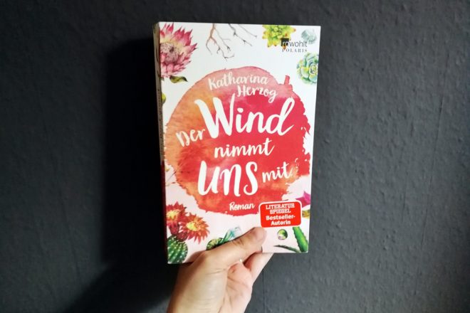 Hand hält das Buch "Der Wind nimmt uns mit" von Katharian Herzog