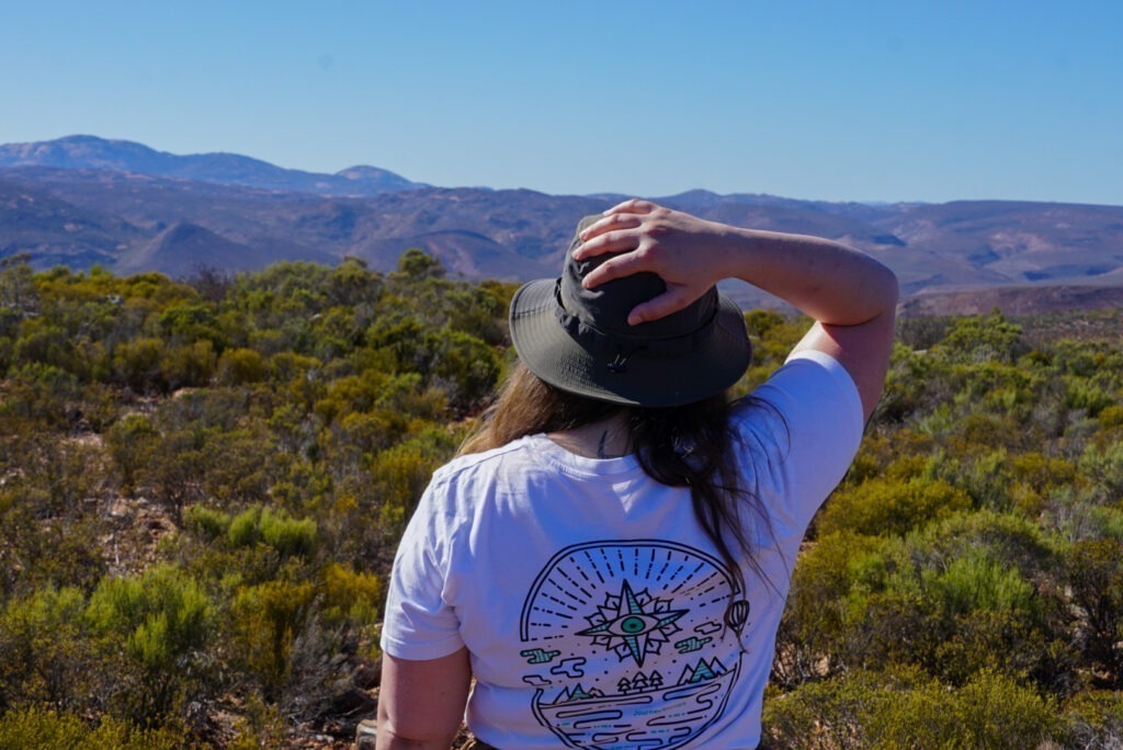 Frau die von hinten fotografiert wurde und ihren Hut hält. Sie guckt in die Weiten der Landschaft des Namaqualands.