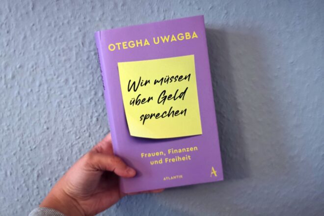 Hand die das Buch "Wir müssen über Geld sprechen" gegen eine grüne Wand hält