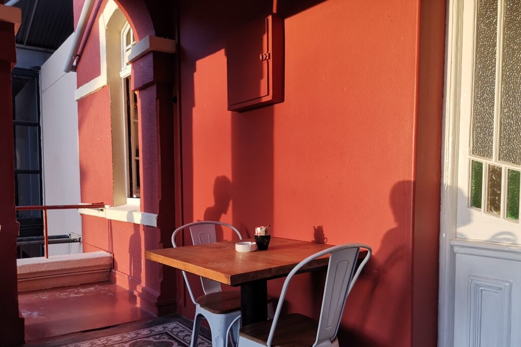 Schatten von Menschen werden auf eine rote Wand in einem Café geworfen. Es sieht beinahe aus, als würden sie auf den Stühlen die vor der Wand stehen, sitzen.
