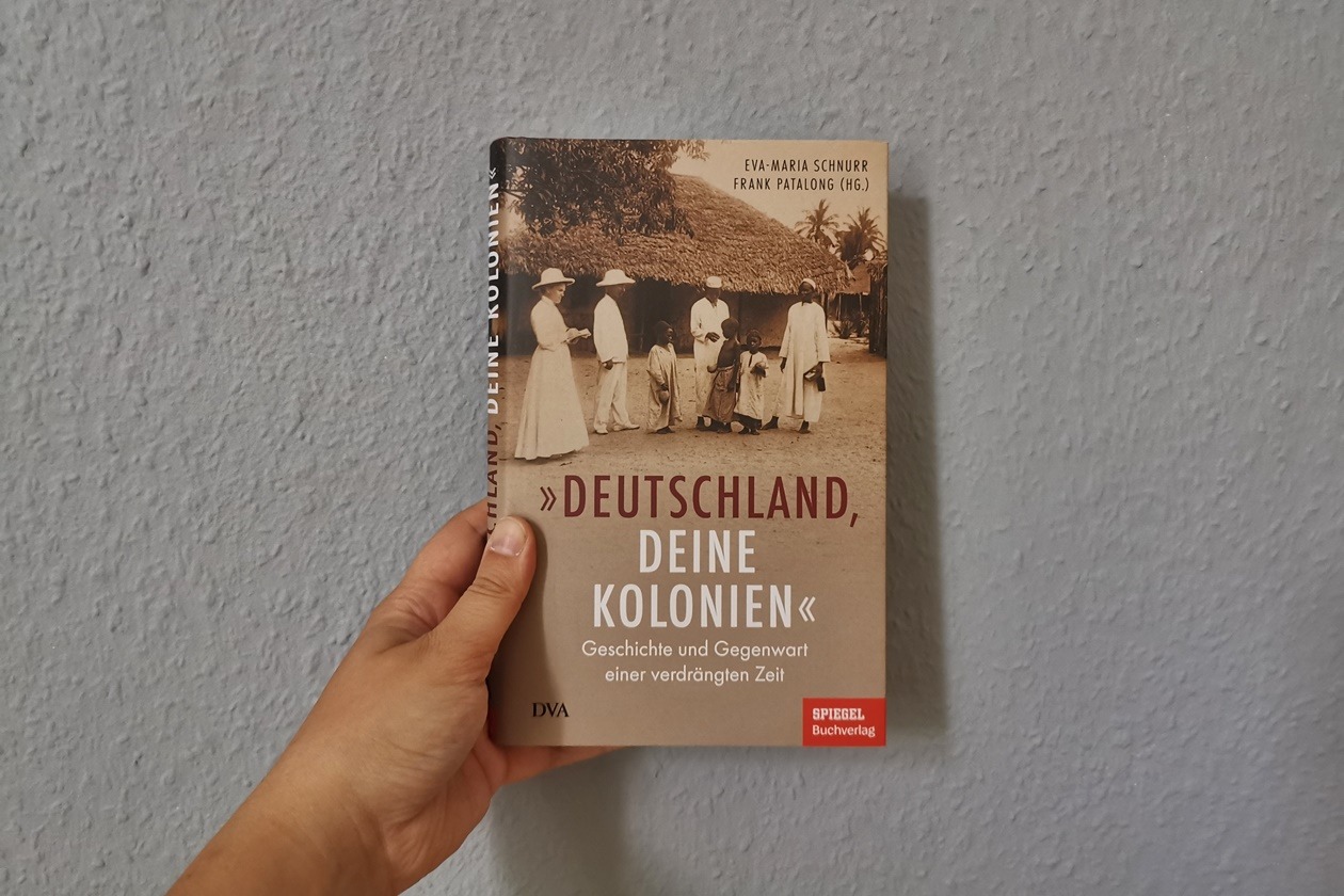 Eine Hand hält das Buch "Deutschland, deine Kolonien" gegen eine hellblaue Hand
