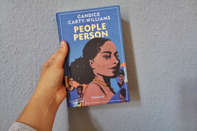 Eine Hand hält den Roman People Person von Candice Carty-Williams gegen eine hellblaue Hand
