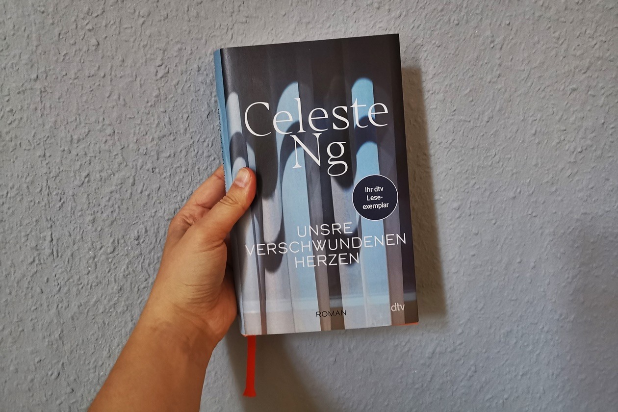 Eine Hand hält den Roman "Unsre verschwundenen Herzen" von Celest Ng gegen eine hellblaue Wand