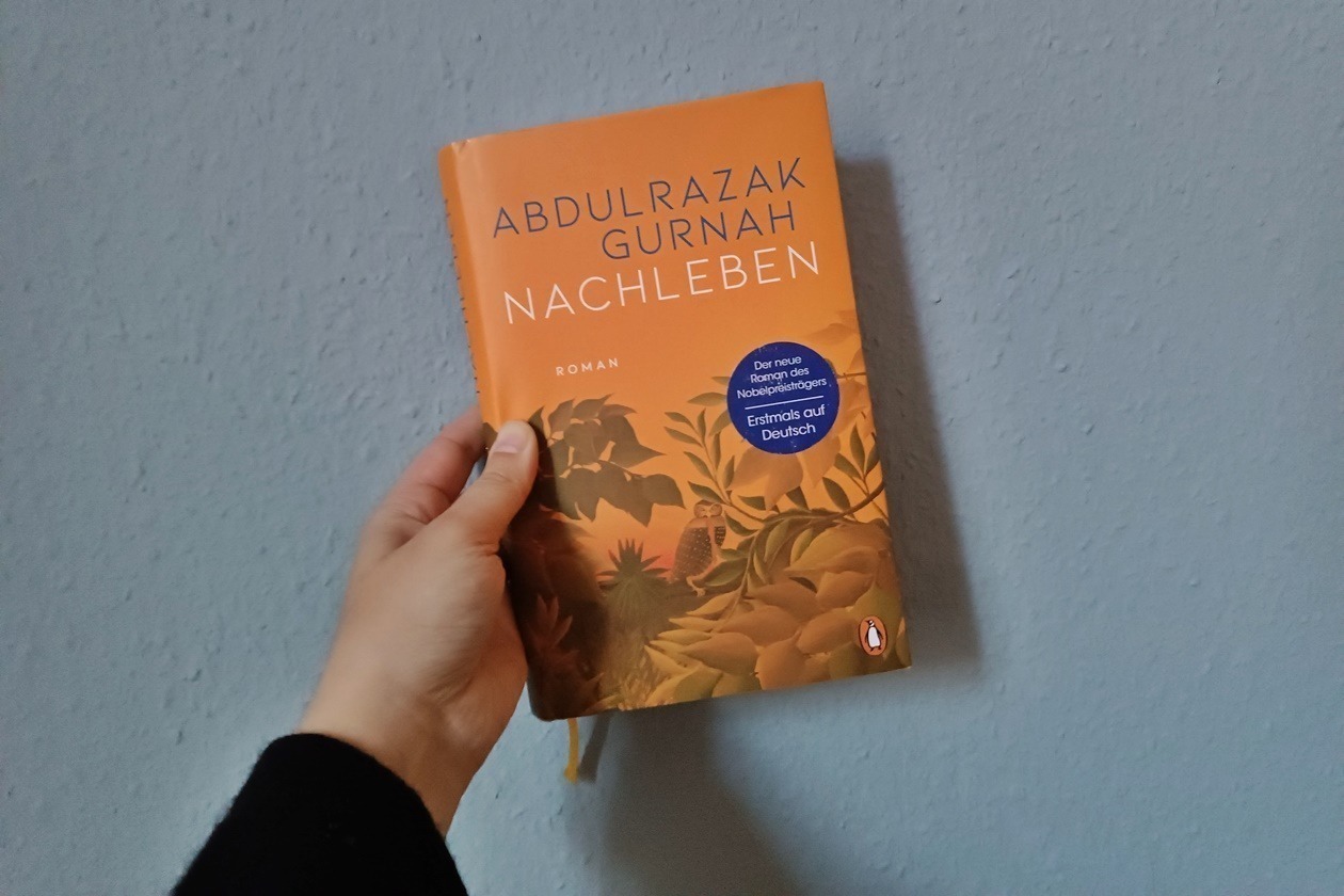 Eine Hand hält den Roman Nachleben von Abdulrazak Gurnah gegen eine hellblaue Hand