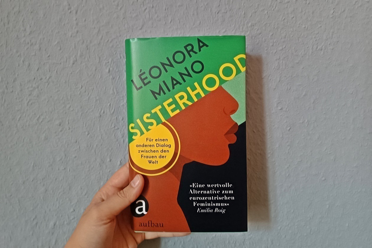 Eine Hand hält das Buch Sisterhood von Léonora Miano gegen eine blaue Wand.