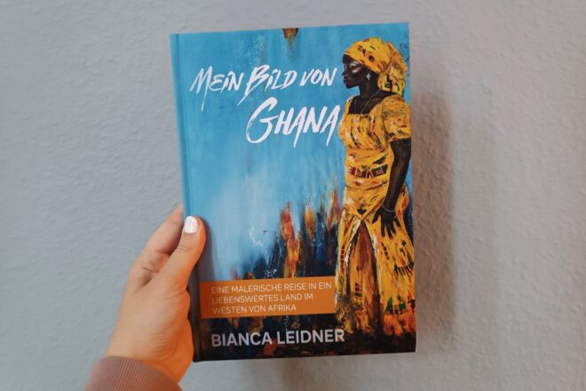 Eine Hand hält das Buch "Mein Bild von Ghana" gegen eine hellblaue Wand.