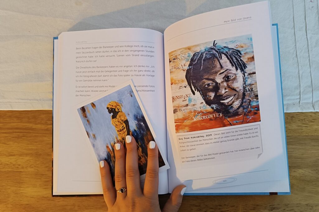 Blick in das Buch "Mein Bild von Ghana". Auf der linken Seite ist bis zur hälfte Text und eine Hand, mit einer Postkarte unter den Fingern, die das Buch offen hält. Auf der rechten Seite ist ein gemaltes Bild von einem lächelnden Mann. Im unteren Drittel ist wieder Text.
