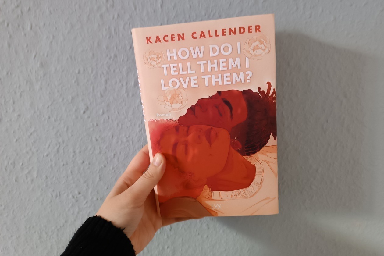 Eine Hand hält das Buch How Do I Tell Them I love Them von Kacen Callender gegen eine hellblaue Wand