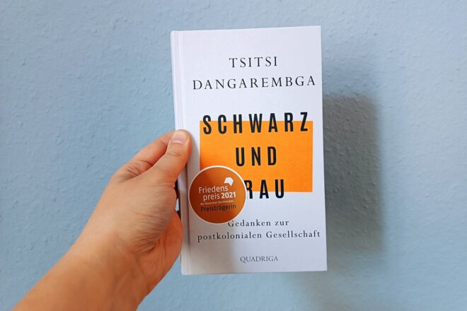 Ein Hand hält das Buch "Schwarz und Frau" von Tsitsi Dangarembga gegen eine hellblaue Wand
