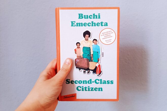 Eine Hand hält das Buch "Second-Class Citizen" von Buchi Emecheta gegen eine hellblaue Wand
