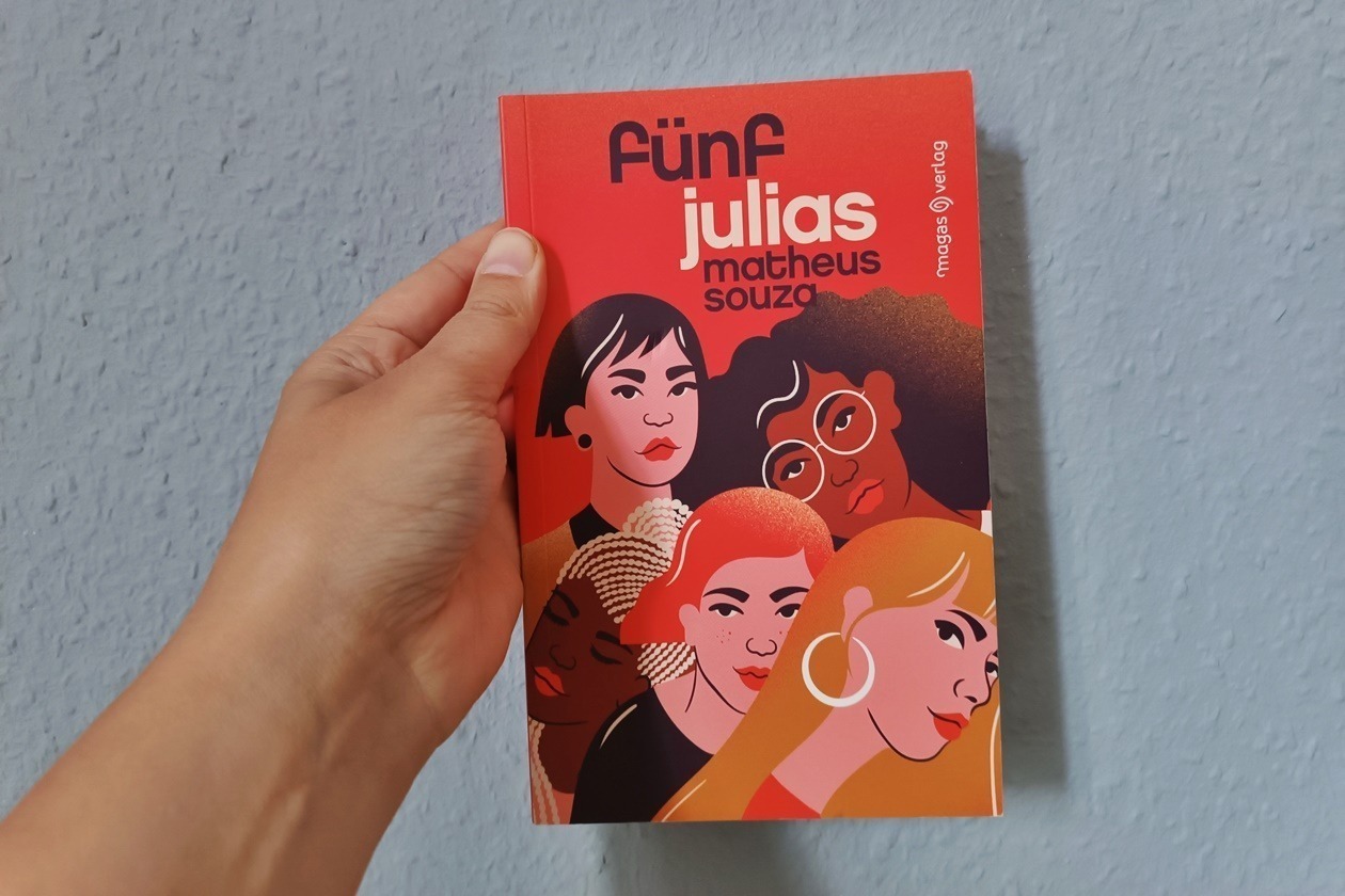 Eine Hand hält das Buch "Fünf Julias" von Matheus Souza gegen eine hellblaue Wand