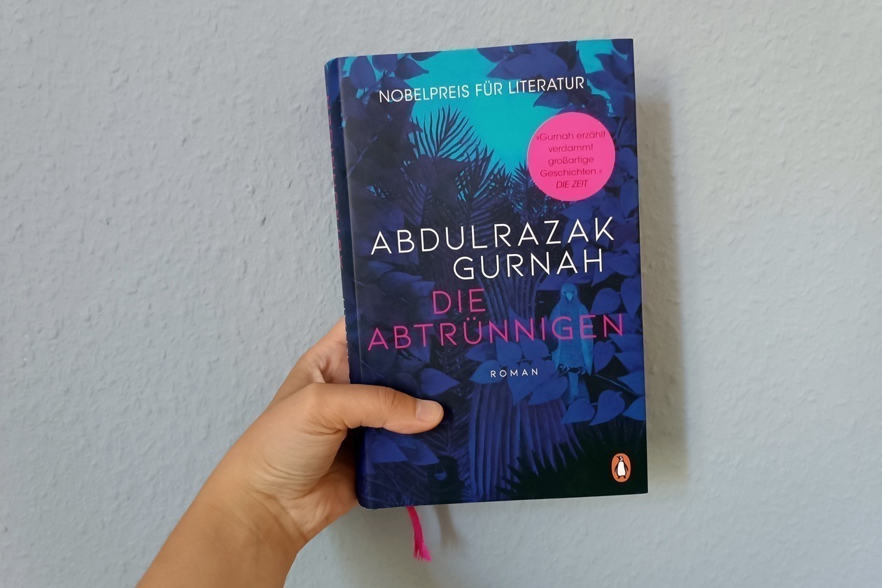 Eine linke Hand hält den Roman Die Abtrünnigen von Abdulrazak Gurnah gegen eine hellblaue Wand.