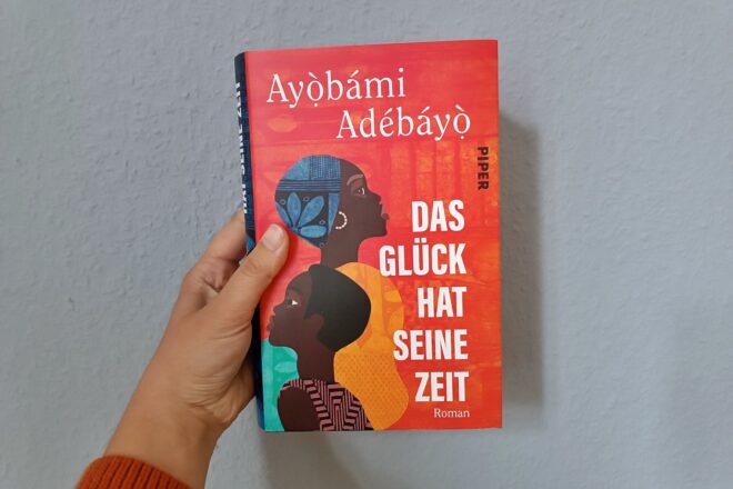 Eine Hand hält den Roman "Das Glück hat seine Zeit" von Ayọ̀bámi Adébáyọ̀ gegen eine hellblaue Wand