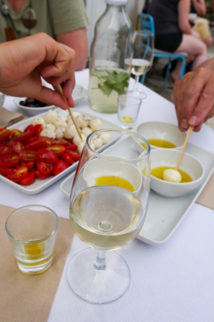 Ein gedeckter Tisch mit Wasser, Wein, Olivenöl, Tomate und Mozaralle. Zwei Hände greifen von jeweils zwei Seiten mit Zahnstochern zu.