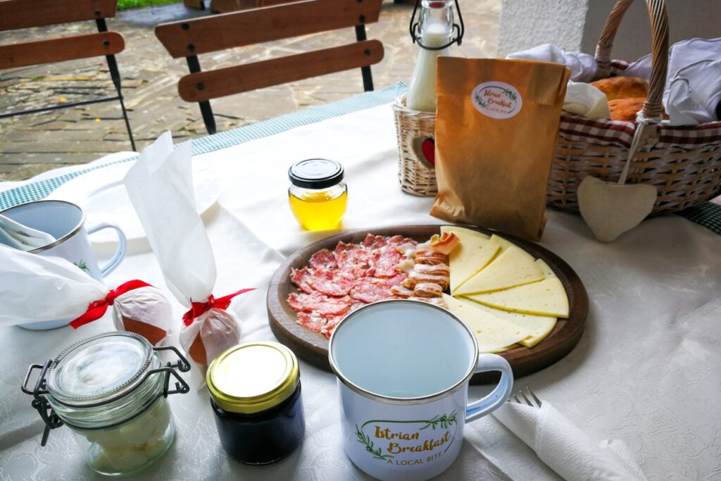 Ein reich gedeckter Frühstückstisch mit slowenischen Spezialitäten. Es gibt Eier, Honig, Marmealde, frische Milch, diverse Salamis und Käse. In der rechten Ecke steht ein Korb mit Brot.