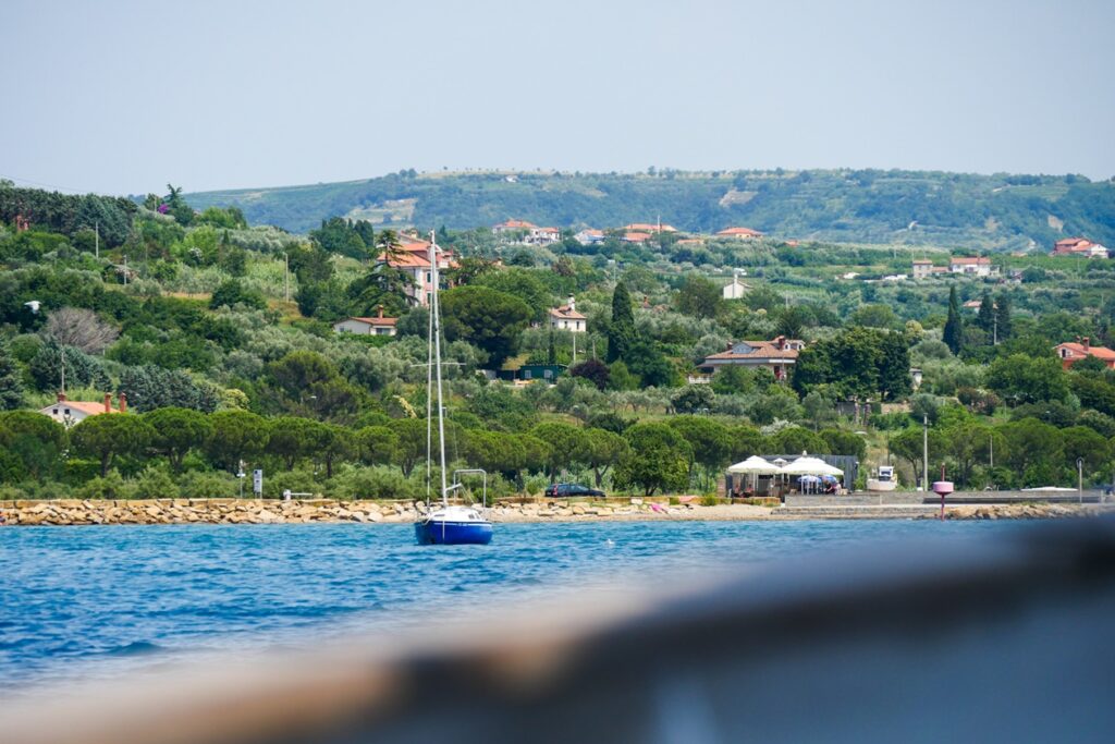Blick über die Rehling eines Bootes auf Sloweniens Mittelmeerküste. Man sieht etwas Meer auf dem ein Boot schaukelt und das begrünte Ufer, wo vereinzelt Häuser stehen.