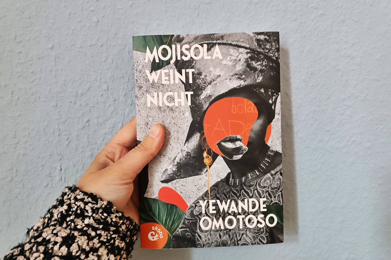 Eine linke Hand hält das Buch "Mojisola weint nicht" von Yewande Omotoso gegen eine hellblaue Wand.