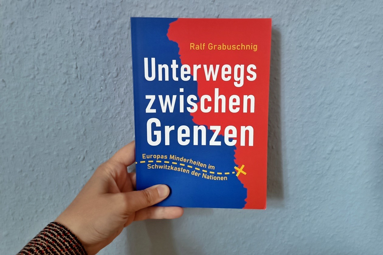 Eine Hand hält das Buch Unterwegs zwischen Grenzen von Ralf Grabuschnig gegen eine hellblaue Wand.