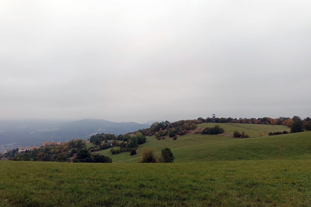 Grüne Hügel unter grauem Himmel. In der Ferne sieht man einen kleinen Aussichtsturm.