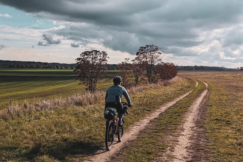 Eine Radfahrerin fährt über einen Feldweg. Am Himmel ist ein Band mit schwarzen Wolken zu sehen.
