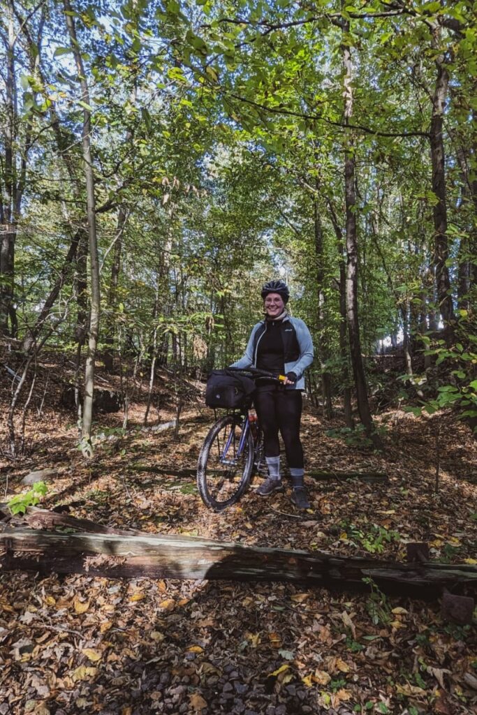 Radfahrerin steht recht von ihrem Rad und lächelt. Der Radweg ist auf einer alten Bahntrasse im Wald und man sieht noch die hölzernen Bohlen.