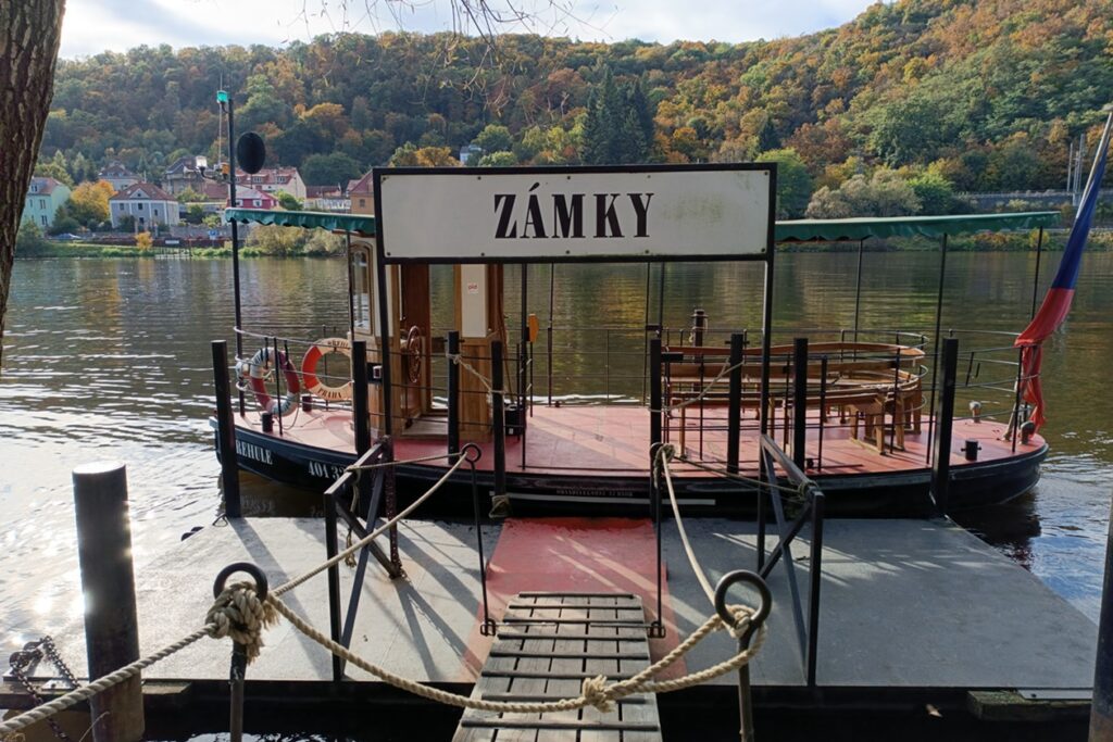 Kleines Holzboot, welches Fußgänger und Radfahrer über die Moldau befördert. Das Boot liegt am Anleger. Darüber ist ein Schild mit der Aufschrift Zamky.