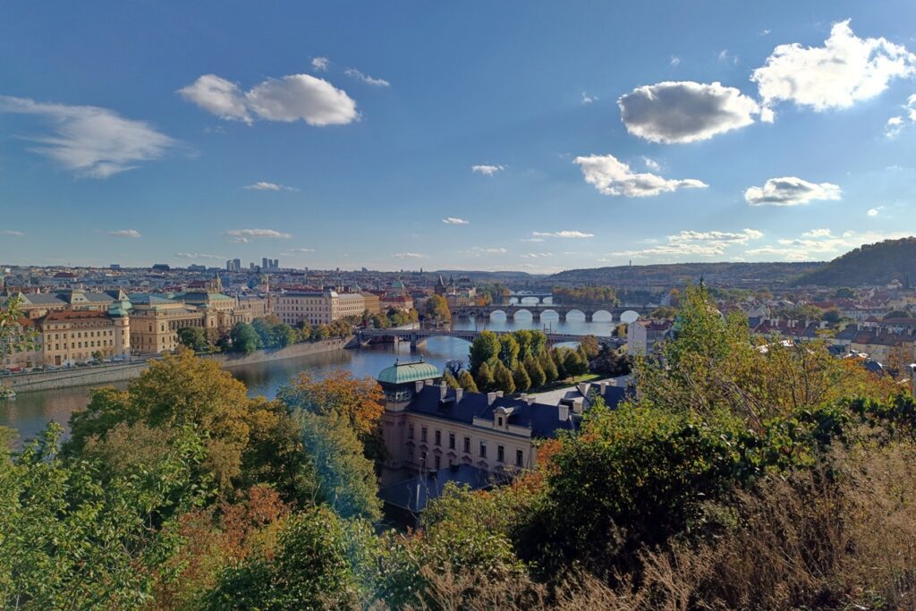 Aussichtspunkt in Prag, von welchem man die Moldau, einige Brücken wie die Karlsbrücke, und die Altstadt überblicken kann. Der Himmel ist blau mit vereinzelten Schäfchenwolken und die Bäume herbstlich gefärbt.
