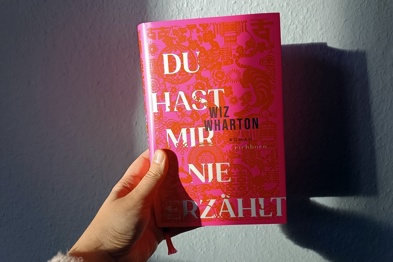 Eine Hand hält das Buch "Du hast mir nie erzählt" von Wiz Wharton gegen eine hellblaue Wand. Die Sonne wirft ein Schattenspiel auf Wand und Buch.