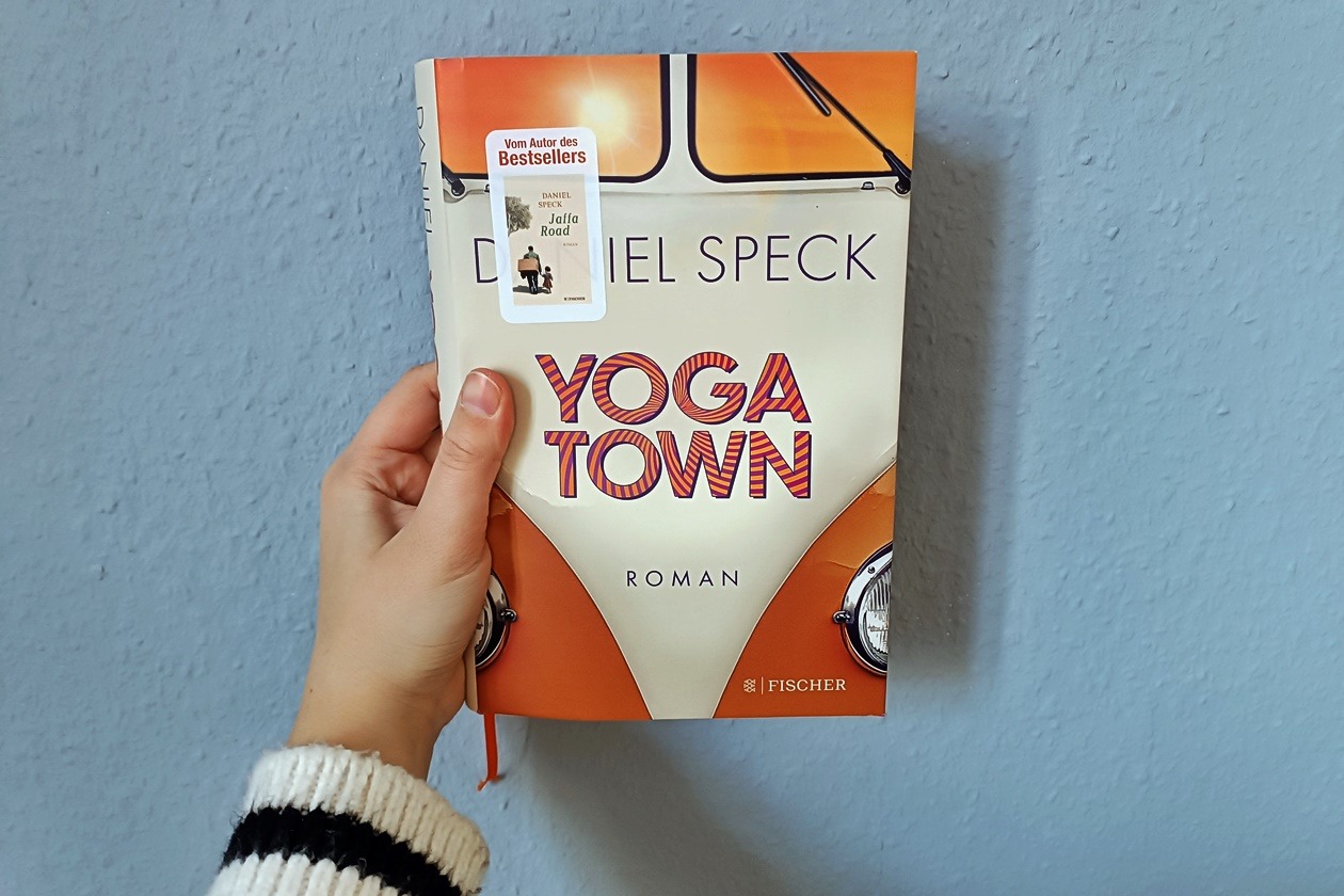 Eine Hand hält das Buch "Yoga Town" von Daniel Speck gegen eine hellblaue Wand.
