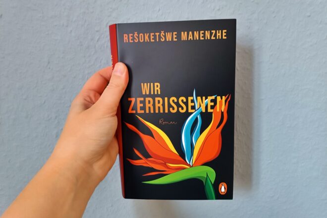 Eine Hand hält das Buch "Wir Zerrissenen" von Rešoketšwe Manenzhe gegen eine hellblaue Wand.
