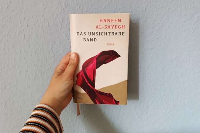 Eine Hand hält das Buch "Das Unsichtabre Band" von Haneen Al-Sayegh gegen eine hellblaue Wand.