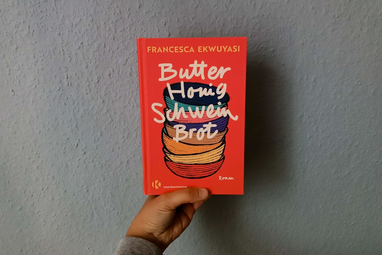 Eine Hand hält das Buch Butter Honig Schwein Brot von Francesxa Ekqwuyasi gegen eine hellblaue Wand.