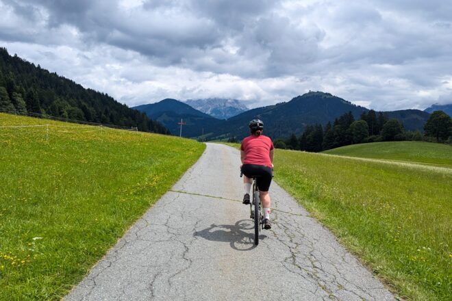 Eine Frau in einem roten Trikot radelt auf dem Radurlaub in St. Johann in Tirol einem Bergpanorama endgegen.