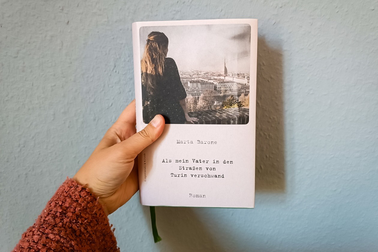 Eine Hand hält das Buch "Als mein Vater in den Straßen von Turin verschwand" von Marta Barone gegen eine hellblaue Wand.