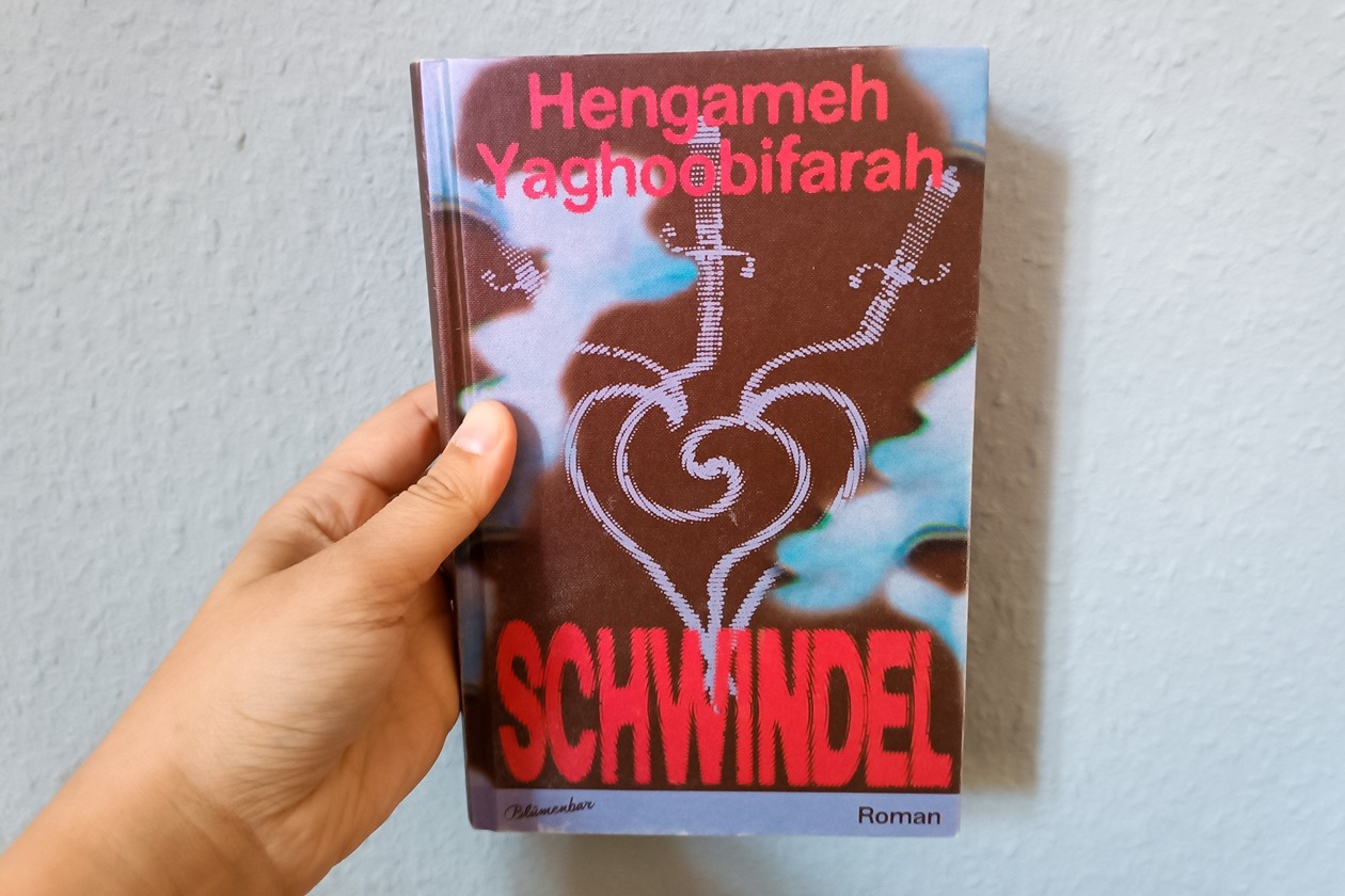 Eine Hand hält das Buch Schwindel von Hengameh Yaghoobifarah gegen eine hellblaue Wand.