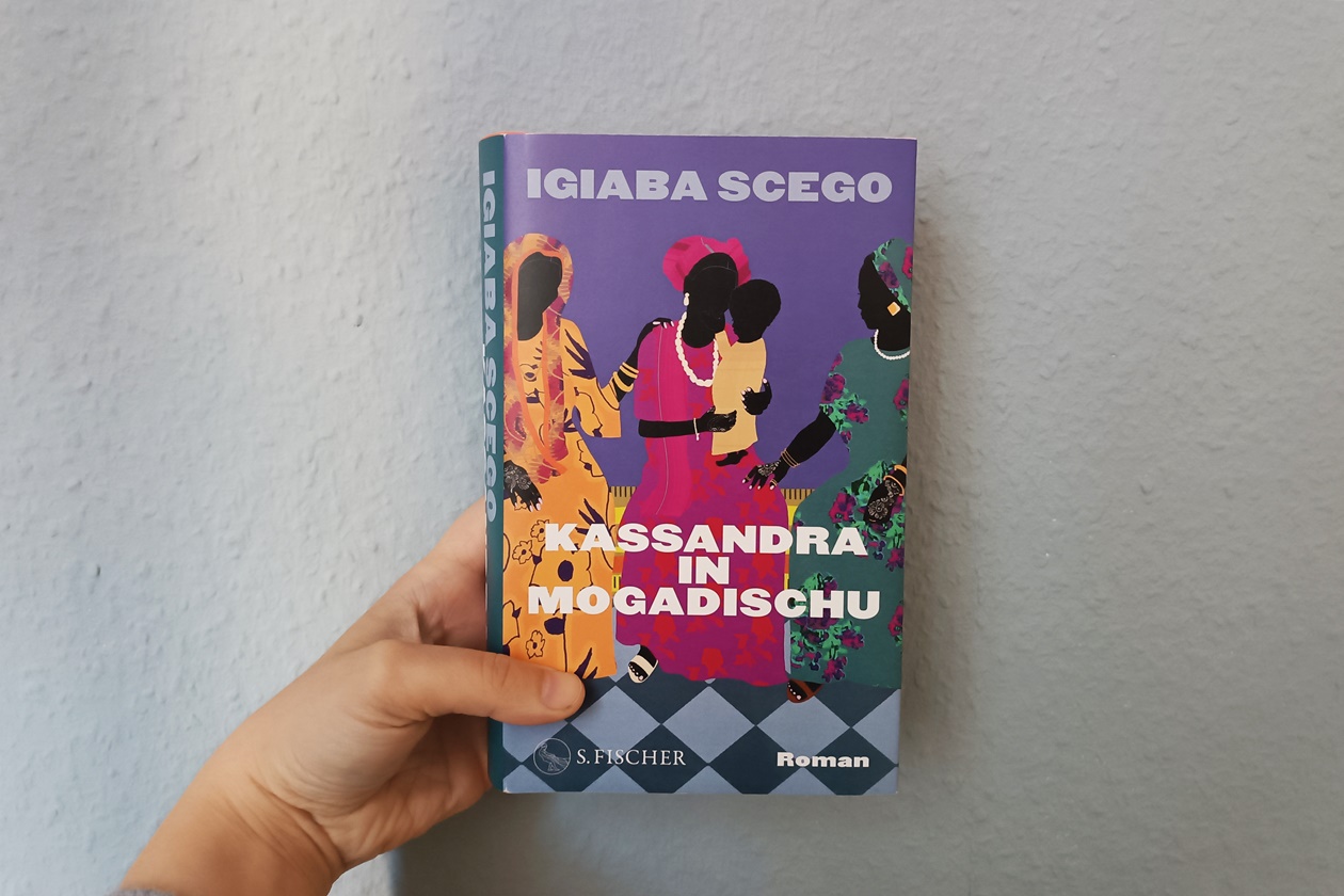 Eine linke Hand hält das Buch "Kassandra in Mogadischu" von Igiaba Scego gegen eine blaue Wand.