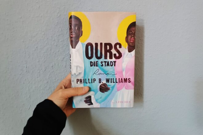 Eine Hand hält den Roman "Ours. Die Stadt" von Phillip B. Williams gegen eine hellblaue Wand.