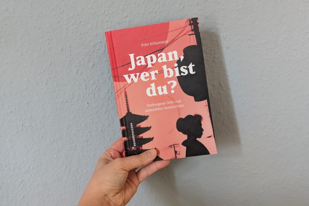 Eine Hand hält das Buch "Japan, wer bist du? gegen eine hellblaue Wand.