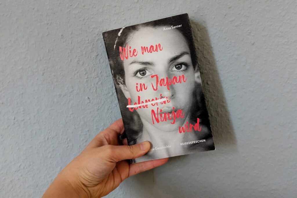 Eine Hand hält das Buch "Wie man in Japan Ninja wird" gegen eine hellblaue Wand.