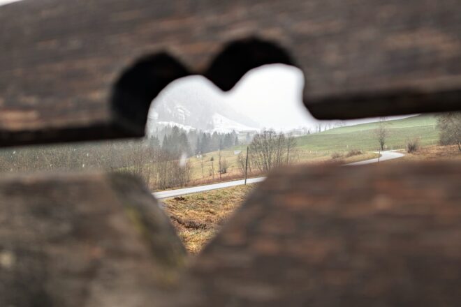 Eine Bank im PillerseeTal, in welchem ein Herz eingeschnitzt ist, durch welche die winterliche Bergwelt fotografiert ist.
