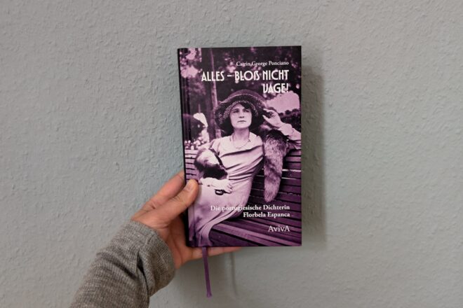Eine Hand mit grauem Ärmel hält das Buch "Alles - bloß nicht vage!" von Catrin George Ponciano über die portugiesische Dichterin Florbela Espanca gegen eine hellblaue Wand.