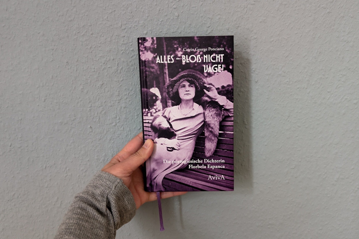 Eine Hand mit grauem Ärmel hält das Buch "Alles - bloß nicht vage!" von Catrin George Ponciano über die portugiesische Dichterin Florbela Espanca gegen eine hellblaue Wand.