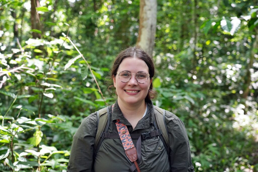 Lynn blick in die Kamera und lächelt. Ihre braunen Haare trägt sie zum Pferdeschwanz gebunden. Im Hintergrund sieht man dichten Wald. Sie trägt einen Rucksack, einen bunten Kameragurt und ein dunkelgrünes Hemd.