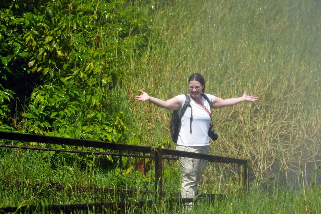 Lynn steht in der Gischt der Murchison Falls. Sie hebt die Arme. Lynn trägt ein weißes T-Shirt, eine Beige Hose, einen Rucksack, eine Kamera mit Gurt über die Schulter und die braunen Haare zum Pferdeschwanz.