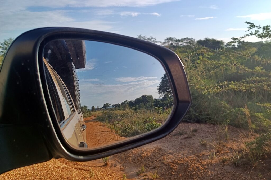 Blick in den Außenspiegel unseres Mietwagens für Uganda. Zu sehen sind Teile des Autos, der blaue Himmel und die rote Straße.