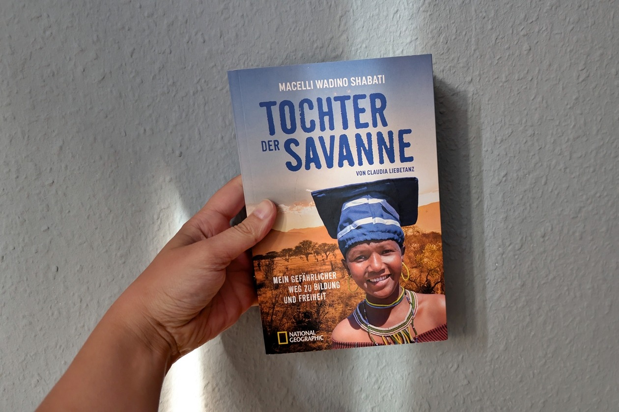 Eine Hand hält das Buch "Tochter der Savanne" von Macelli Wadino Shabati gegen eine hellblaue Wand auf welche Schatten fallen.