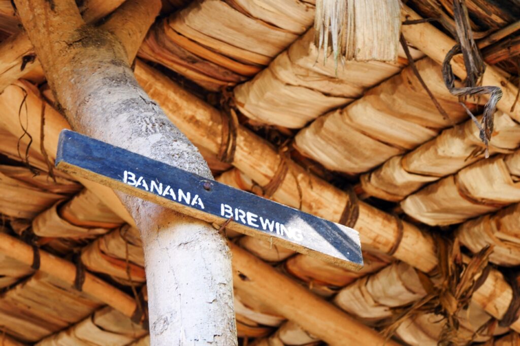Holzschild mit der Aufschrift 'BANANA BREWING' an einem Baumstamm unter einem Dach aus Palmblättern