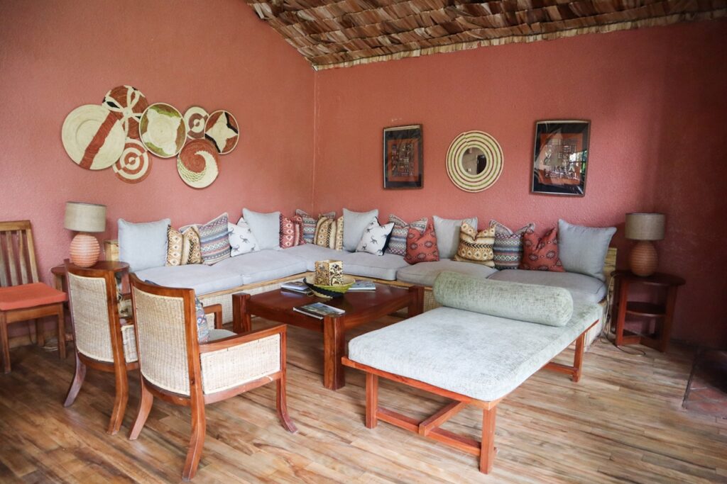 Wohnzimmer mit roter Wand im Mahogany Lodge in Buhoma, Holzboden, Ecksofa mit gemusterten Kissen, zwei Holzstühle, zwei Tischlampen und Wanddekoration aus Körben und Bildern
