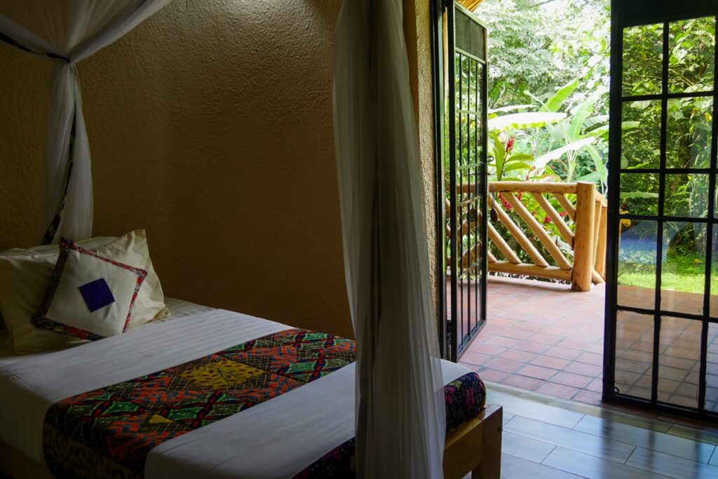 Bett mit Moskitonetz, bunter Decke und Kissen in einem Zimmer von Ride 4 a Woman in Buhoma, offene Glastür führt zu Terrasse mit Holzgeländer und grünem Garten