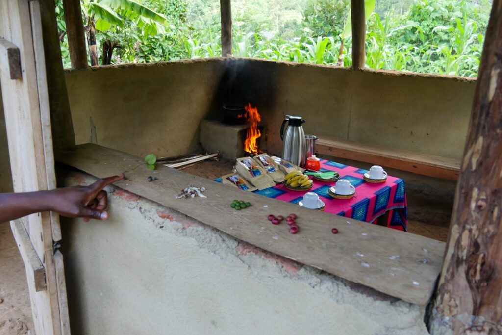 Offene Feuerstelle in einer einfachen Hütte mit brennendem Holz, daneben ein Tisch mit buntem Tuch, Thermoskanne, Tassen und Obst, im Vordergrund eine ausgestreckte Hand